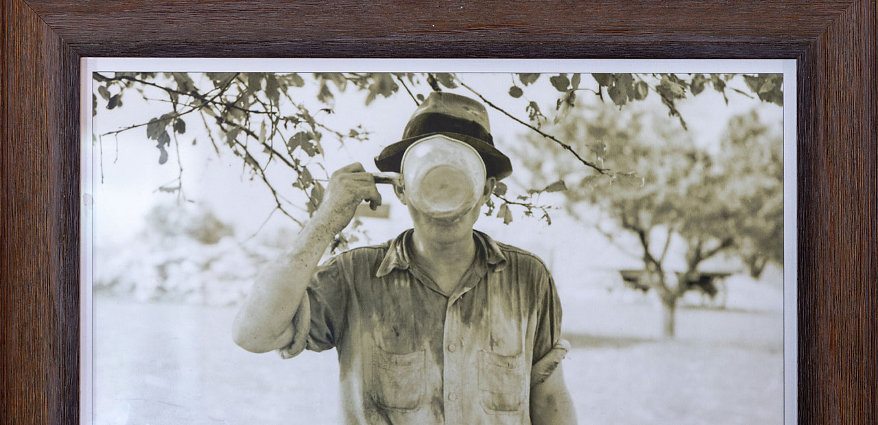 An antique framed photo of a farmer sipping water from a cup in the orchard